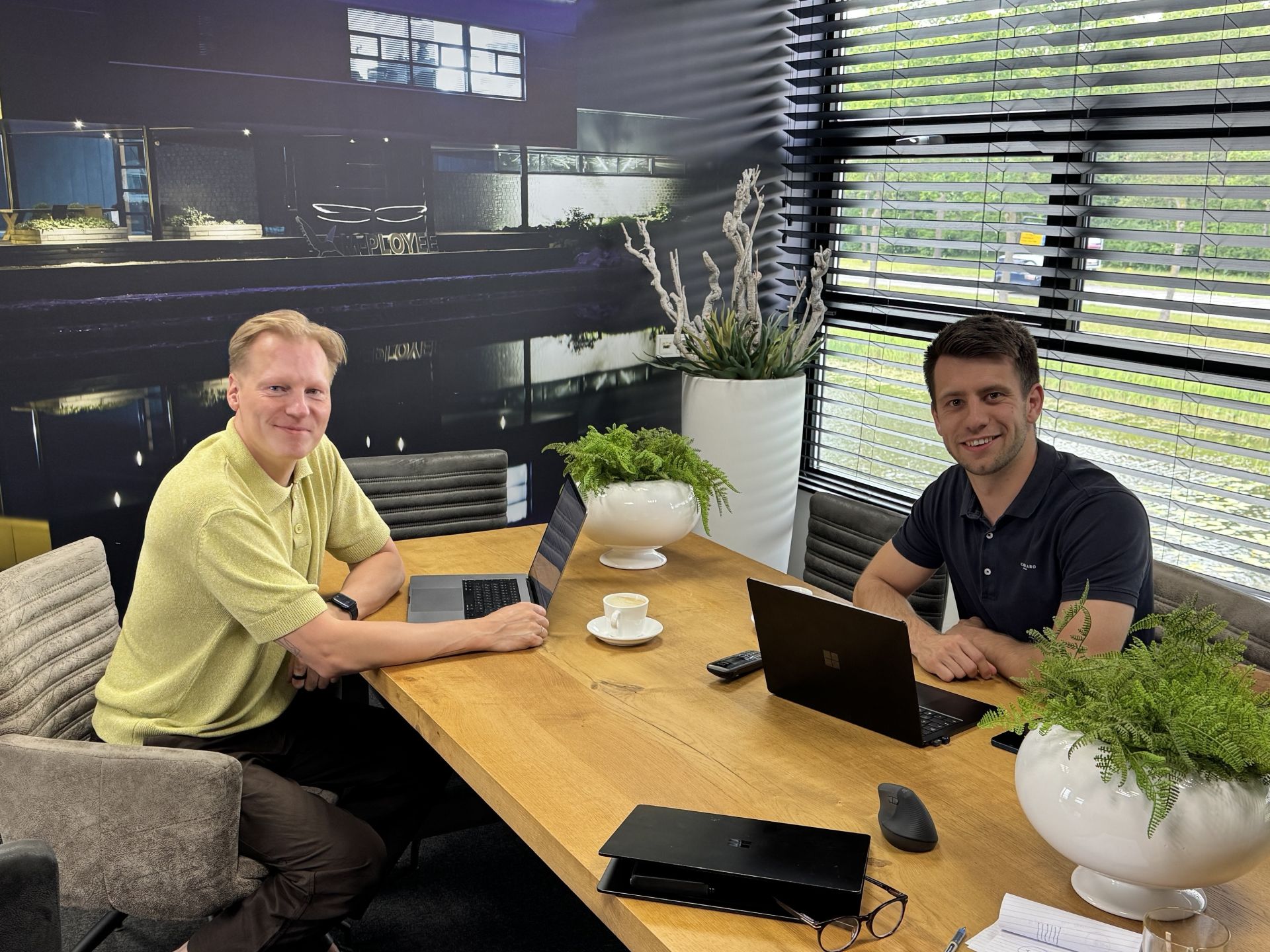 Twee mannen werken op laptops aan een tafel in een kantooromgeving.